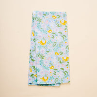 The Hoppy Days Tea Towel by The Darling Effect features light blue fabric with cheerful yellow birds, pink flowers, green leaves, and blue accents, shown folded on a simple beige background.