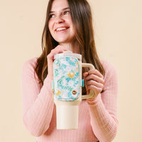 A smiling young woman in a pink sweater holds The Darling Effect’s Hoppy Days Tumbler Fanny Pack—a floral-patterned, water-resistant fanny pack attached to a beige tumbler with straw and sturdy handle. Light beige background.