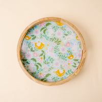 The Hoppy Days Large Round Wood Enamel Tray by the-darling-effect features a colorful, whimsical interior with yellow birds, green leaves, and pink flowers on a light background atop a pale beige surface.