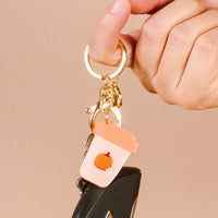 A hand holds The Darling Effect’s Pumpkin Spice Latte Acrylic Keychain—a small, pastel pink cup-shaped charm with an orange pumpkin—attached to keys against a beige background, ideal for cute keychain lovers.