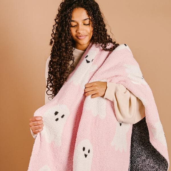 A young woman with curly hair smiles as she hugs her ultra-soft Boo Crew Cozy Dreams Luxe Blanket from The Darling Effect, a soft pink throw featuring white ghosts.