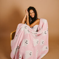 A smiling woman with curly hair sits on a chair, wrapped in The Darling Effect’s Boo Crew Cozy Dreams Luxe Blanket—pink microfiber with playful white ghosts—against a plain beige background.