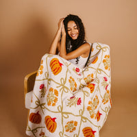 A woman with curly hair smiles while wrapped in The Darling Effect’s Patchwork Pumpkins Cozy Dreams Luxe Blanket—white with orange pumpkins, yellow leaves, and red flowers—against a beige background.