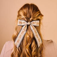 A person with wavy, reddish-blonde hair styled half up, adorned with The Darling Effect’s Cream Cottage Crush Hair Bow featuring a floral-patterned ribbon. The background is a soft peach color.