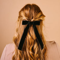 A person with wavy, reddish-blonde hair styled half-up, wearing The Darling Effect's Black Vintage Velvet Hair Bow, is photographed from the back against a plain beige background.