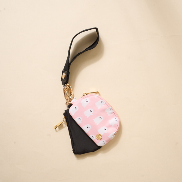 The Darling Effect's Boo Crew Dynamic Duo Pouch Wristlet features a black and pink design with white ghosts, a luxe gold zipper, and a black wrist strap—all stylishly arranged on a beige surface.