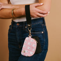 A person in blue jeans and a white top wears The Darling Effect’s Boo Crew Dynamic Duo Pouch Wristlet, a black wrist strap attached to a pink ghost-pattern pouch for carrying essentials hands-free.