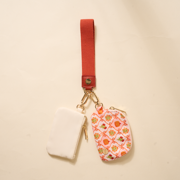 The Darling Effect’s Patchwork Pumpkins Dynamic Duo Pouch Wristlet pairs two zip pouches—one solid white, one pink floral—on a luxe red strap with gold hardware for a stylish, hands-free accessory.