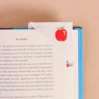 A blue hardcover book lies open with The Darling Effect’s Oh So Orchard Corner Keeper Fabric Bookmark, featuring a red apple design, resting on the top corner—a perfect gift for book lovers set against a light beige background.