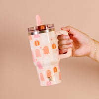 A hand holds The Darling Effect's Ghost Party 20 oz Lifestyle Flip Straw Tumbler, featuring a handle, pink straw and lid, and cute pastel ghosts and pumpkins on a beige background—perfect for fall sipping on the go.