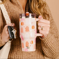 A person in a brown sweater holds The Darling Effect Ghost Party 20 oz Lifestyle Flip Straw Tumbler, featuring ghosts and pumpkins. They also carry car keys and a white bag—perfect for fall outings with their favorite fall tumbler.