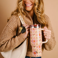 A smiling woman in a tan sweater holds The Darling Effect Patchwork Pumpkins 40 oz Lifestyle Flip Straw Tumbler and a white purse with car keys attached, against a solid beige background.