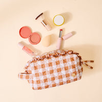 The Maple Gingham Essentials Quilted Cosmetic Pouch by The Darling Effect, adorned with a charming checkered pattern, is displayed on a light beige background surrounded by blush, lip gloss, a brush, blending sponge, and lipstick. Featuring a wipeable interior, this cosmetic bag effortlessly blends style with functionality.