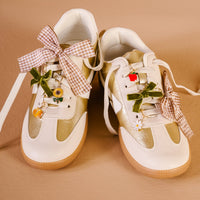 The Darling Effect’s Hayride Honey Dazzle Shoe Charm Pack features gingham and green velvet bows, playful sneaker charms—like flowers, a red apple, and a yellow fruit—that add standout style to white and beige sneakers with brown soles.