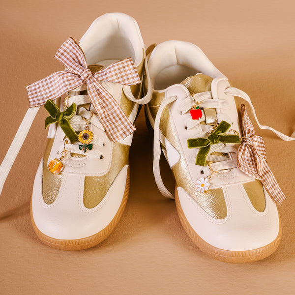 The Darling Effect’s Hayride Honey Dazzle Shoe Charm Pack features gingham and green velvet bows, playful sneaker charms—like flowers, a red apple, and a yellow fruit—that add standout style to white and beige sneakers with brown soles.