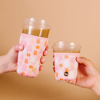 Two hands hold iced coffees with The Darling Effect’s Ghost Party Sip Savvy Drink Sleeves—one with a pink ghost, one with a pumpkin. Against a light brown backdrop, the stylish neoprene sleeves pop. One cup is clearly larger than the other.