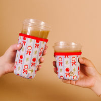 Two hands hold iced drinks in clear cups, each wrapped in The Darling Effect’s Oh So Orchard Sip Savvy Drink Sleeve featuring red bows, apples, and gold accents on a light background, against a plain beige backdrop.