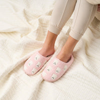 A person wearing light beige pants sits on a cream-colored textured blanket, with their feet in The Darling Effect’s Ghosts Fuzzy Slippers.