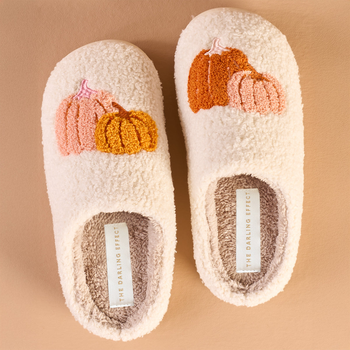 Two cream-colored fuzzy slippers with embroidered orange and pink pumpkins on top, placed side by side on a light brown surface. The insoles are gray and labeled THE DARLING EFFECT..