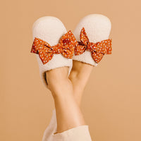 A person relaxes with feet up against a beige background, wearing The Darling Effect's Betty Bow Fall Flora Fuzzy Slippers featuring large red floral bows.