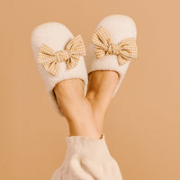 A person with crossed legs in light pants wears The Darling Effect's Betty Bow Maple Gingham Fuzzy Slippers, featuring cozy fall vibes and large checkered bows, set against a beige background.