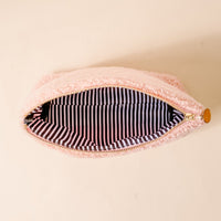 The Darling Effect’s Ghosts & Pearls Teddy Pouch, crafted from cozy pink sherpa with a gold zipper pull, is shown open to reveal its black and white striped lining—a charming fall accessory resting on a beige surface.