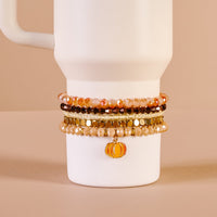 The Darling Effect's Autumn Glow Tumbler Bracelet Stack, featuring orange, gold, and brown beads with a pumpkin charm, decorates a white 40 oz tumbler cup displayed on a beige background.