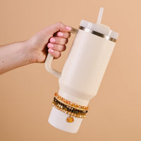 A hand with pink nail polish holds a cream 40 oz tumbler with a straw. The Darling Effect Autumn Glow Tumbler Bracelet Stack adds flair around the handle base, set against a solid beige background.