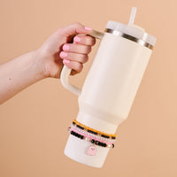 A hand with pink nails holds a 40 oz tumbler featuring The Darling Effect’s Spook & Sparkle Tumbler Bracelet Stack and a cute pink charm on the handle, all set against a solid beige background.