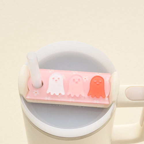 A white straw peeks out from a Stanley Quencher 2.0 lid adorned with The Darling Effect's Ghost Party Tumbler Lid Tag—cute ghost charms that make perfect personalized tumbler accessories, set on a pink floral background.