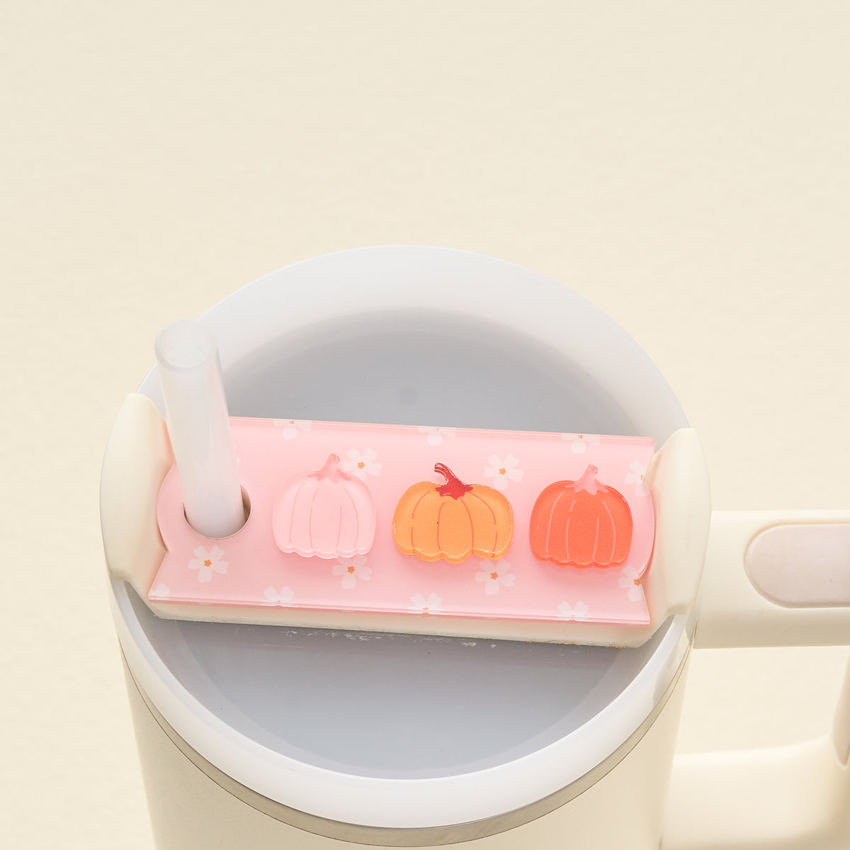 A pink straw cover with three pumpkins and white flowers sits on top of a white cup filled with a grayish liquid. A straw is inserted through the cover.