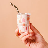 A hand holds The Darling Effect's Ghost Party 2 oz Tiny Tumbler, featuring a cute pastel ghost design on a light background with a metal straw, set against a peach-colored backdrop.