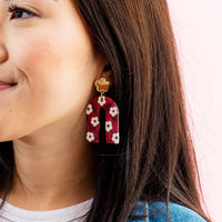 A woman with long dark hair smiles as she wears The Darling Effect's Burgundy Game Day Acrylic Earrings—large, arch-shaped, burgundy with white flowers and a gold stud—perfect for showing off your school colors or on game day.