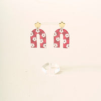 A pair of burgundy, arch-shaped acrylic earrings with white flowers and gold stud tops by The Darling Effect, displayed on a clear acrylic stand—perfect for showing off your school colors.