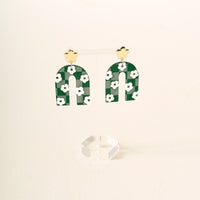 Show off your school colors with the Green Game Day Acrylic Earrings by The Darling Effect—green arch-shaped acrylic earrings with white flowers, hanging from gold flower studs and displayed on a clear stand against a light background.
