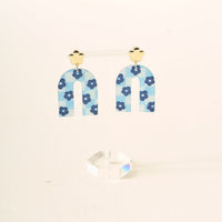 The Darling Effect's Light Blue Game Day Acrylic Earrings feature arch shapes in blue tones with a checkered pattern, small blue flowers, and gold-colored flower studs. Displayed on a clear stand with a white background—perfect for school spirit or game day.