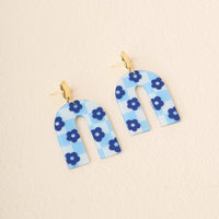 Light Blue Game Day Acrylic Earrings by The Darling Effect feature arched acrylic with a blue and white checkered pattern, blue flower accents, and gold stud posts. Perfect for showing school spirit. Displayed on a light beige surface.