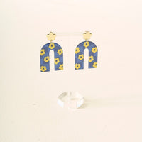 Show off your school colors with The Darling Effect’s Navy Yellow Game Day Acrylic Earrings, featuring gold-colored flower studs and blue arch dangles with yellow flowers, displayed on a clear stand against a light background.