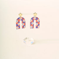 The Darling Effect's Orange Purple Game Day Acrylic Earrings feature gold clover studs and arch dangles in an orange and purple checkered pattern with white flowers, perfect for showing school spirit, displayed on a clear acrylic stand.