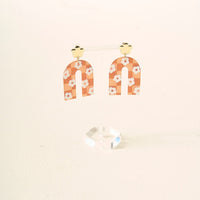The Darling Effect's Orange Game Day Acrylic Earrings feature gold cloud-shaped studs and arched, peach dangles with white daisy patterns, displayed on a clear acrylic stand against a light background.