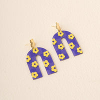 Show your school spirit with The Darling Effect’s Purple Yellow Game Day Acrylic Earrings—featuring a purple arch, yellow flowers, and checkered pattern, all stylishly set against a cream background.