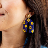 Close-up of a woman wearing The Darling Effect’s Purple Yellow Game Day Acrylic Earrings, featuring a U-shaped purple and yellow checkered design dangling from a gold flower stud—ideal for showing school spirit on game day.