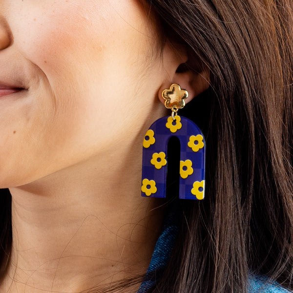 Close-up of a woman wearing The Darling Effect’s Purple Yellow Game Day Acrylic Earrings, featuring a U-shaped purple and yellow checkered design dangling from a gold flower stud—ideal for showing school spirit on game day.