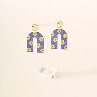 Show your school spirit with The Darling Effect's Purple Yellow Game Day Acrylic Earrings—arch-shaped with yellow flower patterns on a purple background, hanging from gold cloud studs, and displayed on a clear stand.
