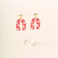 A pair of red, arch-shaped acrylic earrings with white flower patterns hang from gold-colored flower-shaped studs, displayed on a clear stand—Red Game Day Acrylic Earrings by The Darling Effect, perfect for showing your school spirit on game day.