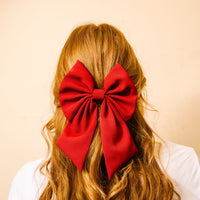 A person with long, wavy, reddish-brown hair is shown from behind, wearing The Darling Effect’s Burgundy Hair Bow as a bright accessory. The background is a solid light beige color.