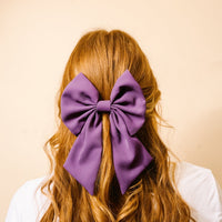 A person with long, wavy, reddish-brown hair is seen from behind wearing The Darling Effect’s Purple Hair Bow. The background is a plain light beige color.