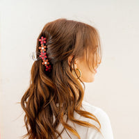 A woman with wavy brown hair, seen from behind and the side, wears a white top, large hoop earrings, and styles her hair with The Darling Effect's Burgundy Game Day Daisy Claw Clip plus a pink-and-red floral hairpin.