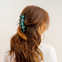 A person with long, wavy brown hair styled half-up wears The Darling Effect's Green Game Day Daisy Claw Clip, featuring blue-green daisy accents in school colors, with a white top, facing away from the camera against a plain background.