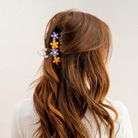 A woman with long, wavy brown hair is seen from behind, wearing The Darling Effect’s Orange Purple Game Day Daisy Claw Clip in her hair, paired with a white top against a plain background—perfect for showing school spirit.